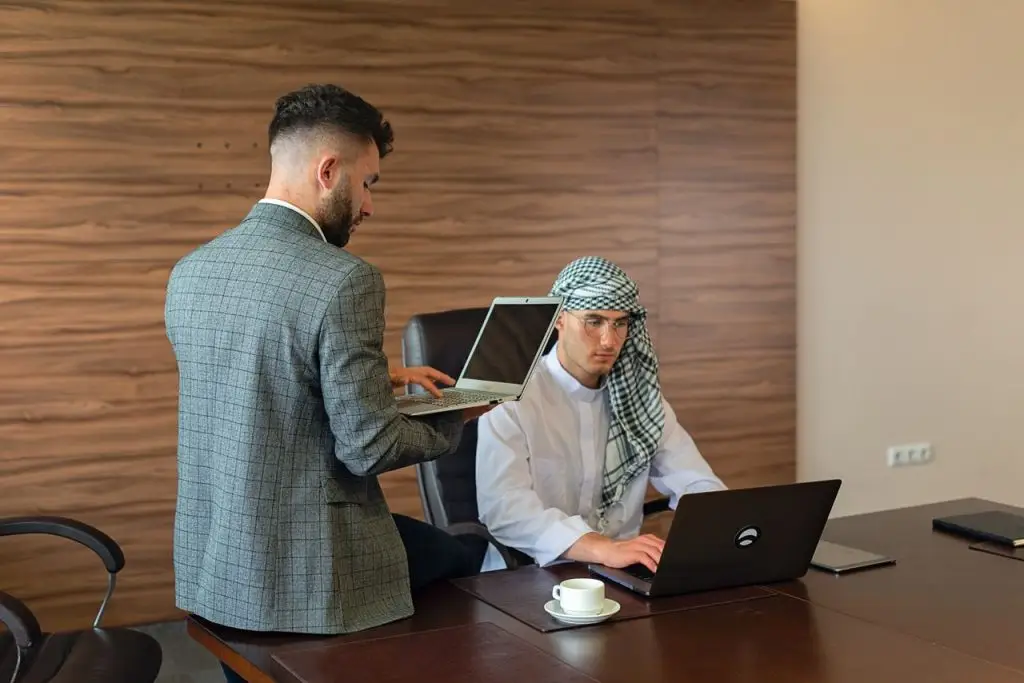 Two men working together in a modern office setting with laptops and a wooden backdrop.